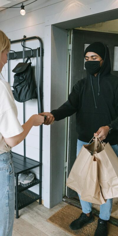 A masked courier hands over shopping bags to a customer in a contactless delivery exchange.