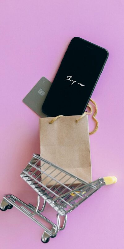 A smartphone, credit card, and bag in a miniature shopping cart on a pink background.